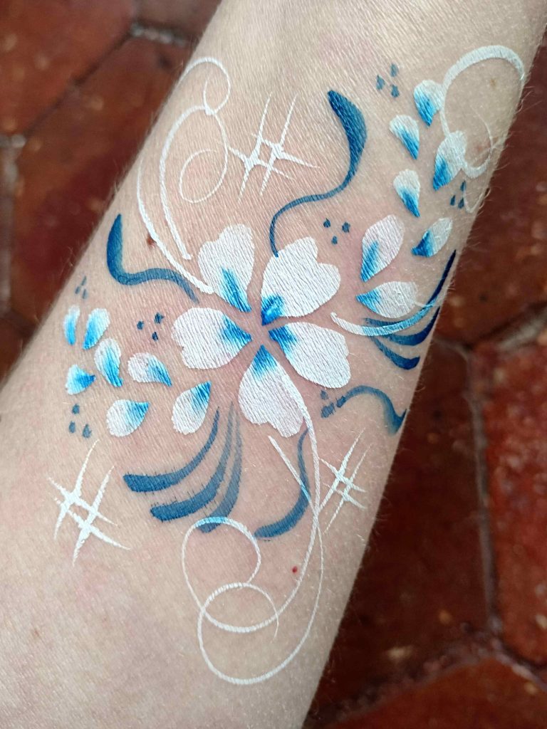 Face-painting sur le bras avec un motif de fleurs bleues avec des pétales et des arabesques par Camille F créations