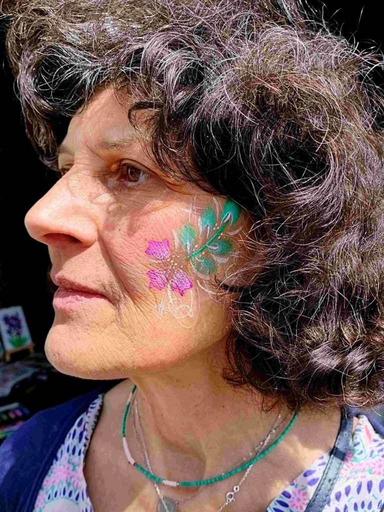 Face-painting de fleur rose esprit hibiscus et feuillage sur la joue par Camille F créations